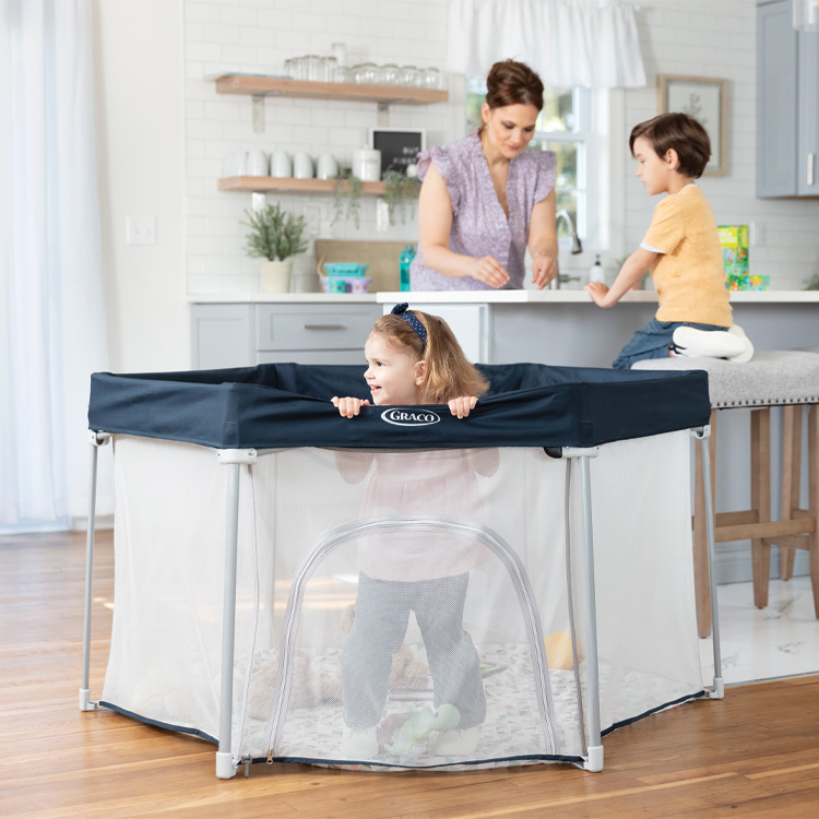 Toddler standing in Graco EverGo playpen with mum and brother at kitchen counter.