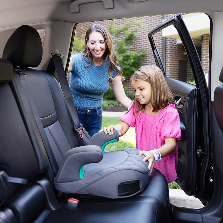 Little girl placing Graco Booster Max R129 ISOFIX backless booster the vehicle while her mum watches behind her
