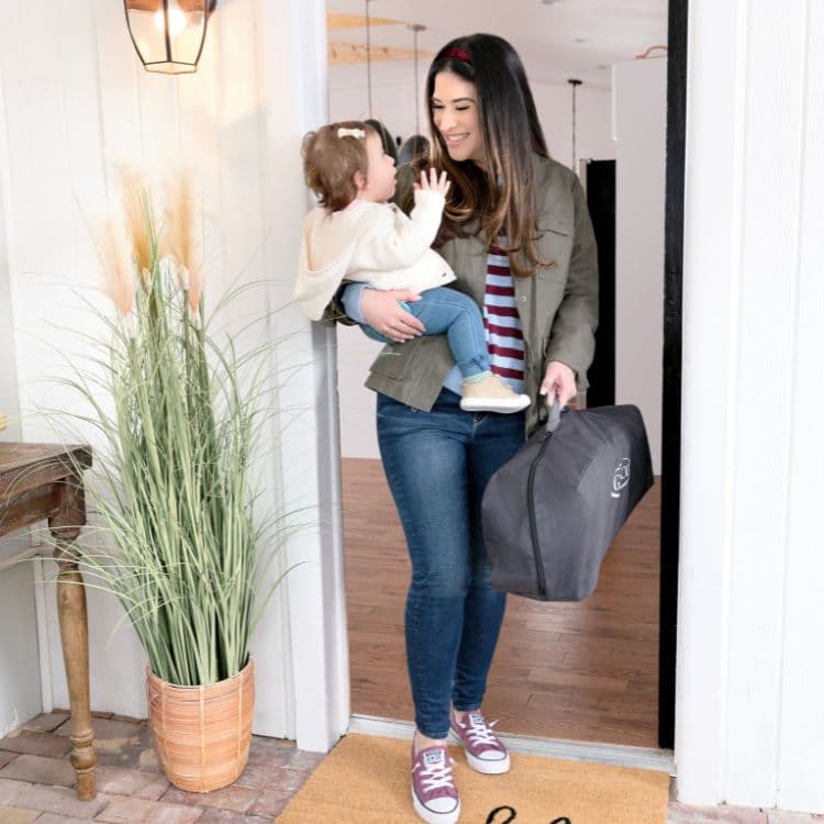 Mum holding toddler and Graco FoldLite™ LX Lightweight Travel Cot in doorway