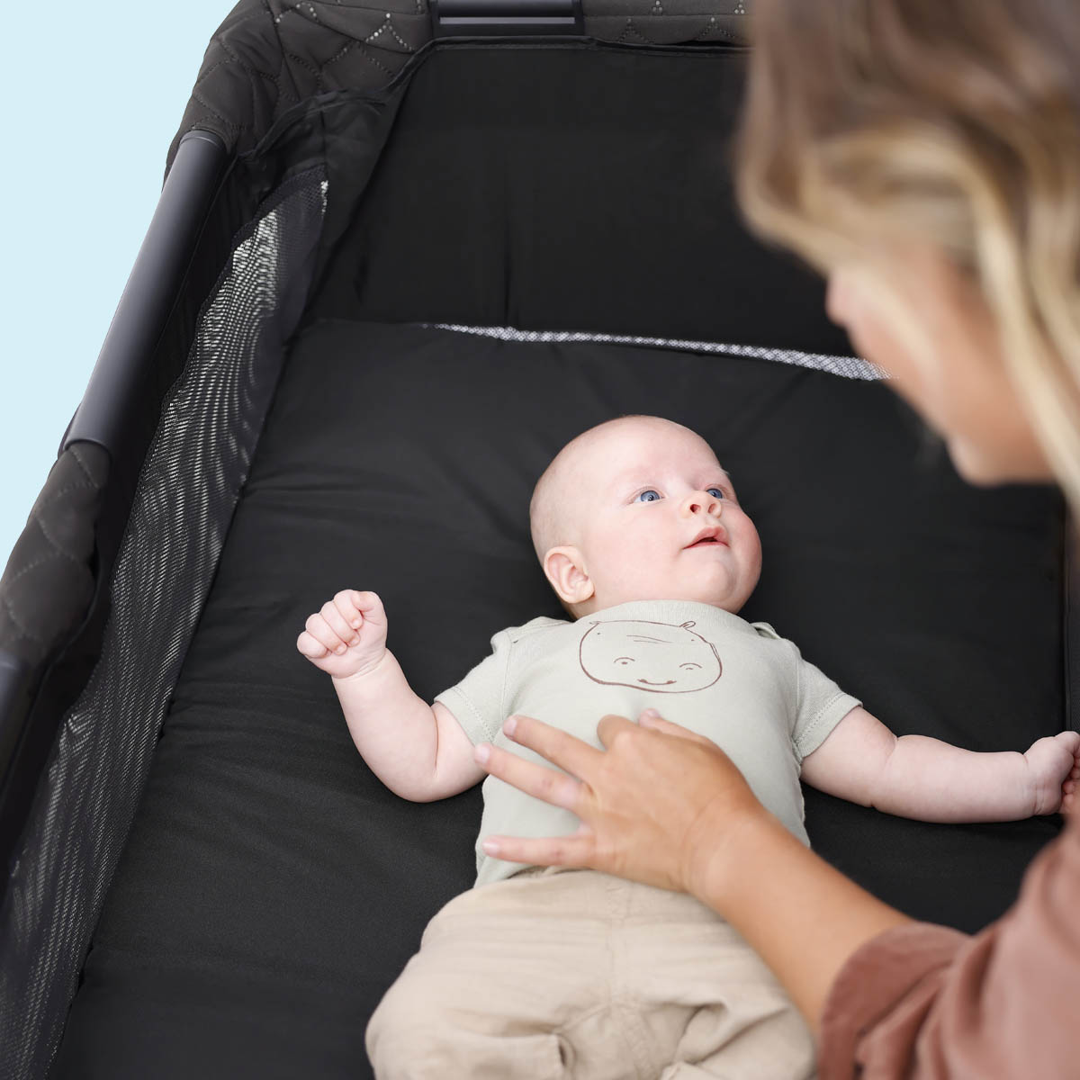 Mutter beschäftigt sich mit Baby, das im Graco FoldLite™ LX Reisebett mit eingehangenem Bassinet-Einhang liegt.
