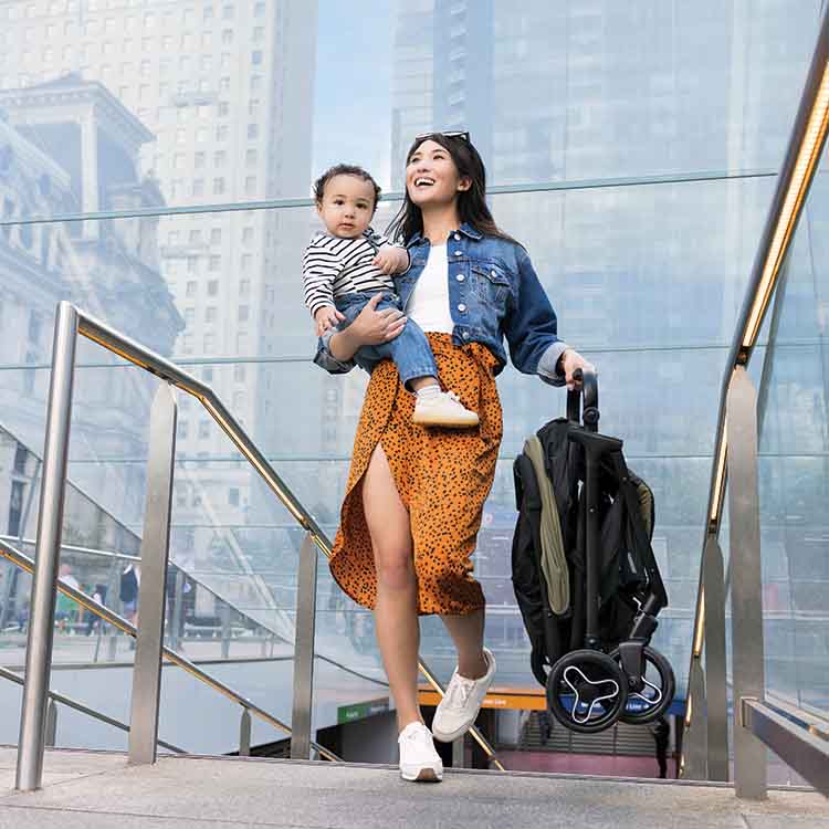 Maman montant les escaliers en portant une poussette de voyage Myavo Graco dans une main et un bébé souriant dans l'autre.