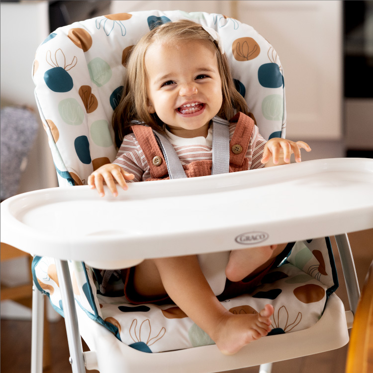 Little girl sitting and smiling in Graco SnackEase highchair with her hands on the removable tray
