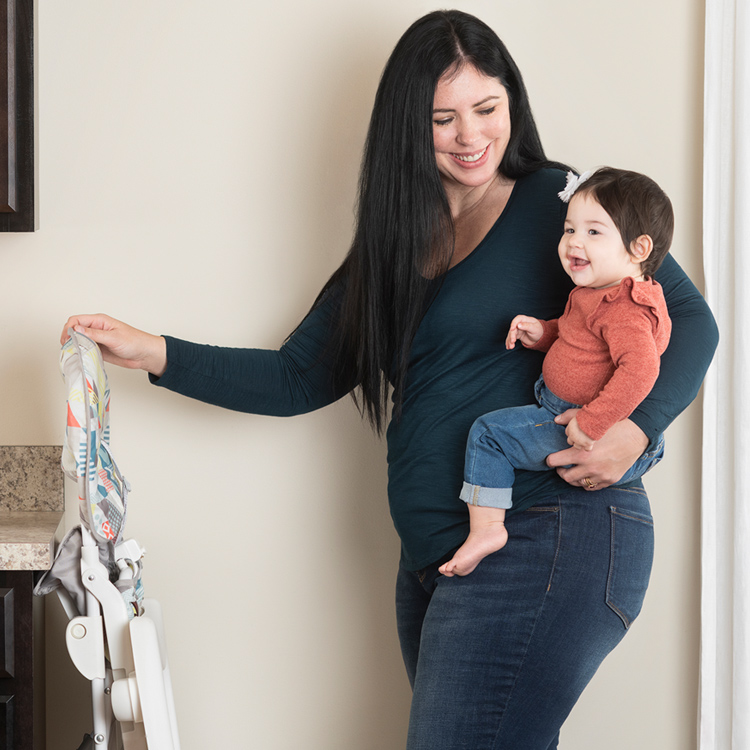 Maman tenant un bébé avec une main pendant qu'elle plie le Graco Snack N' Stow avec l'autre main