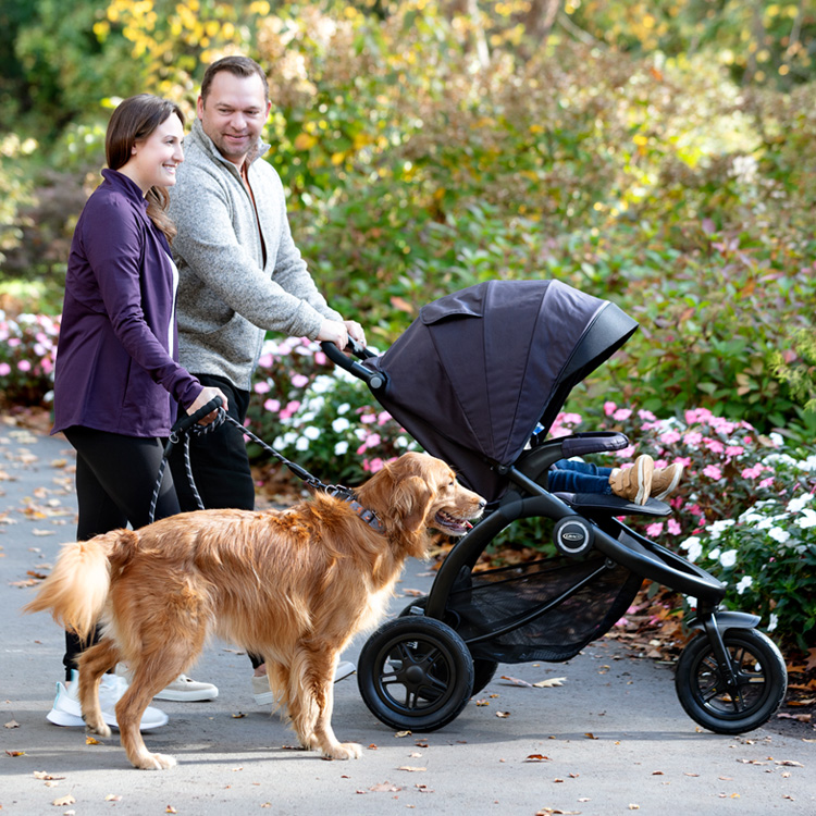 Vater schiebt Kind im 3-Rad-Kinderwagen Graco TrailRider, während Mutter einen Hund an der Leine ausführt